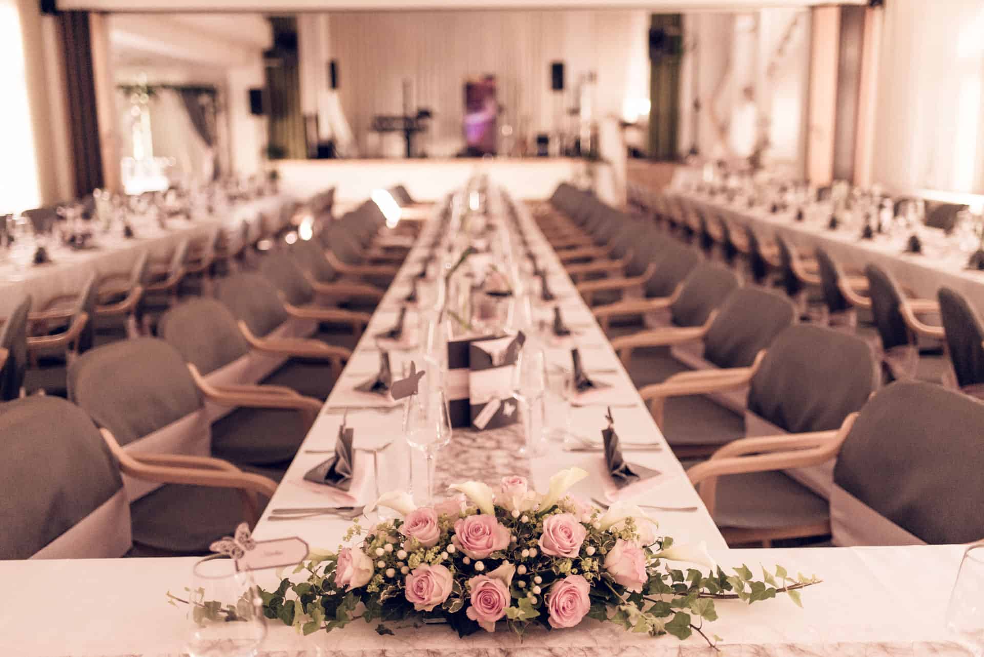 Hochzeitssaal - Ein langer Tisch mit weißen Tischdecken und rosa Blumen dekoriert